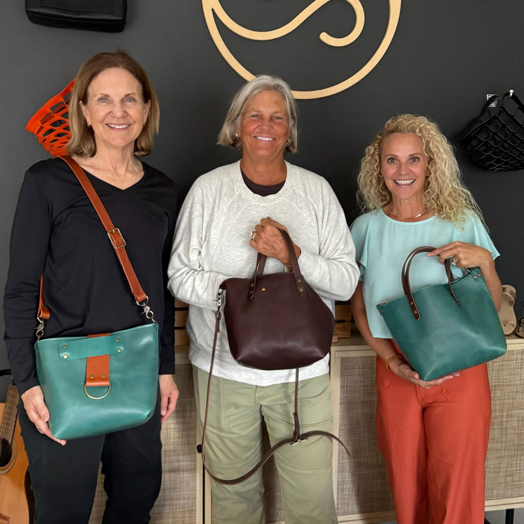 Three women holding handbags in a store setting with a decorative wall in the background.