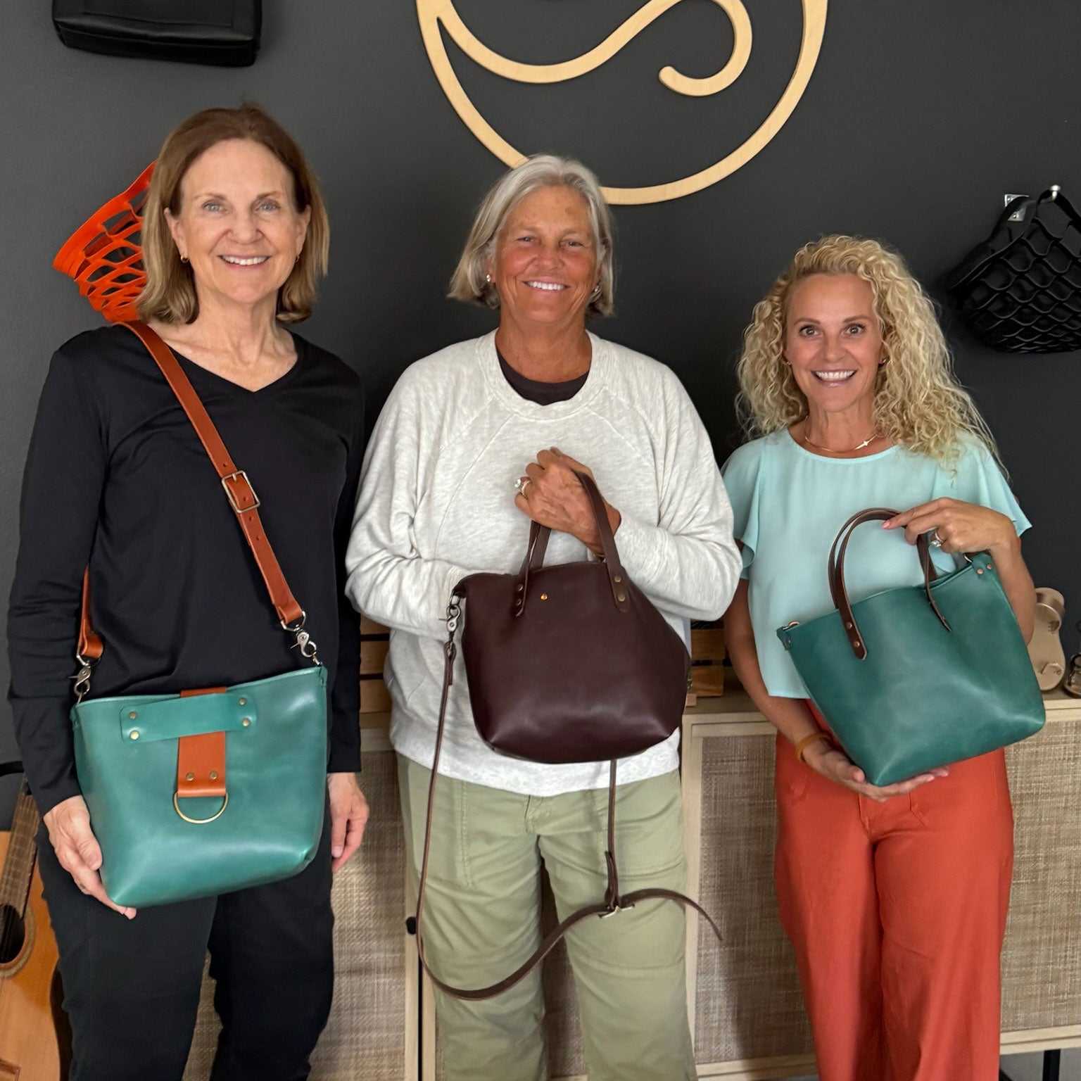 Three women holding handbags in a store setting with a decorative wall in the background.