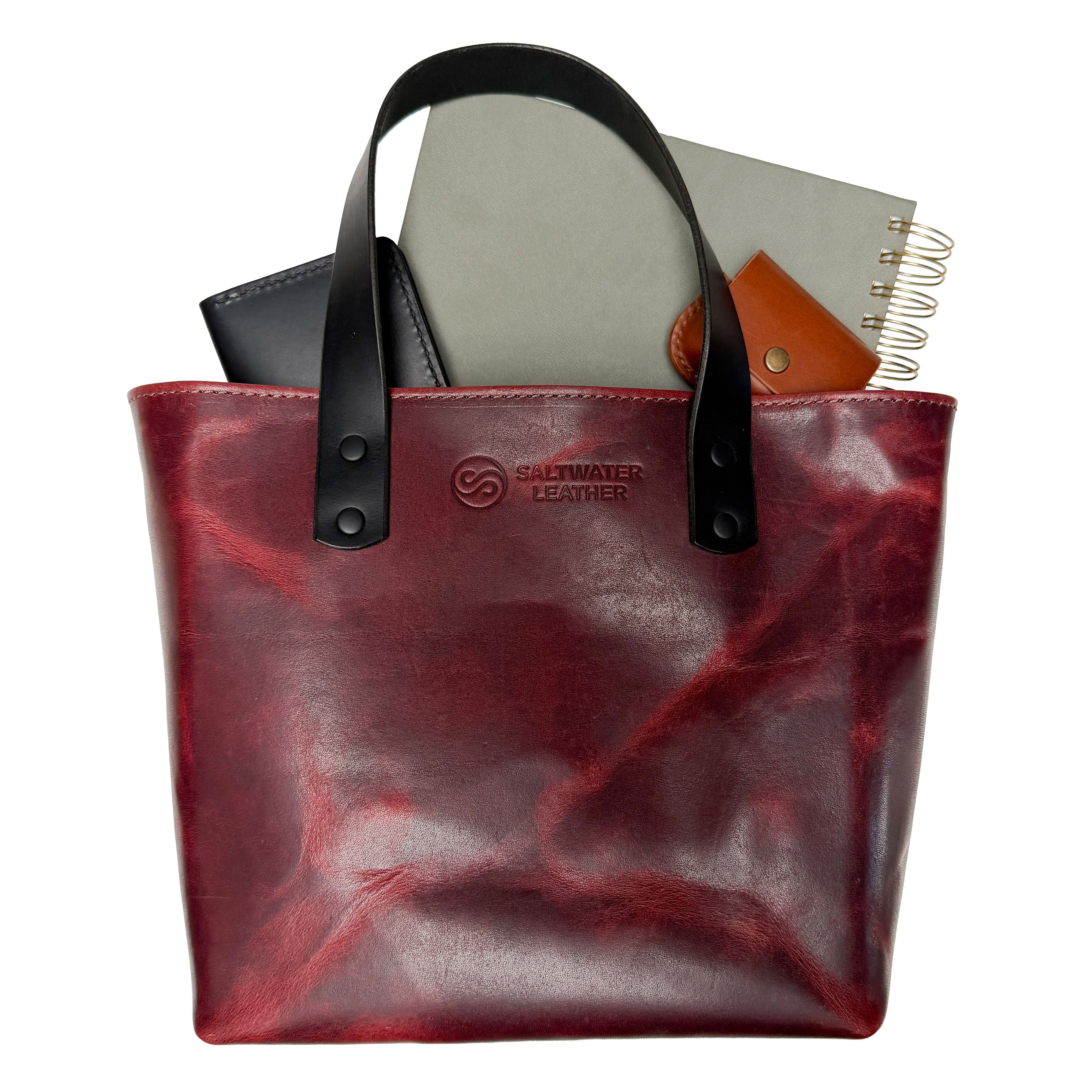 Red leather tote bag with black handles and Saltwater Leather logo on a white background. Green notebook, brown airpod case, and black wallet are inside the bag.