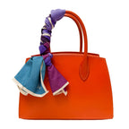 Orange handbag with a purple and blue scarf tied to it on a white background