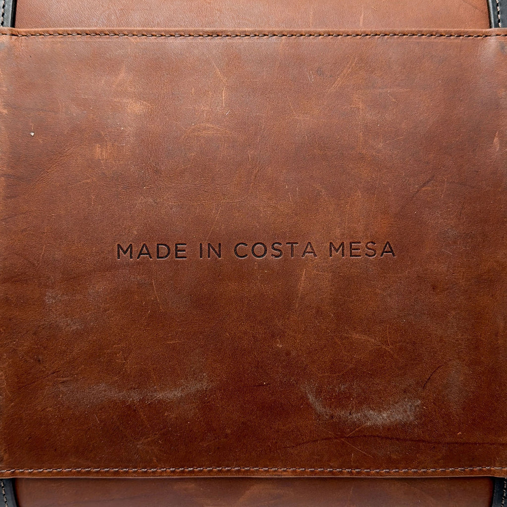 Brown leather duffle bag with MADE IN COSTA MESA text embossed on it.