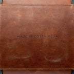 Brown leather duffle bag with MADE IN COSTA MESA text embossed on it.