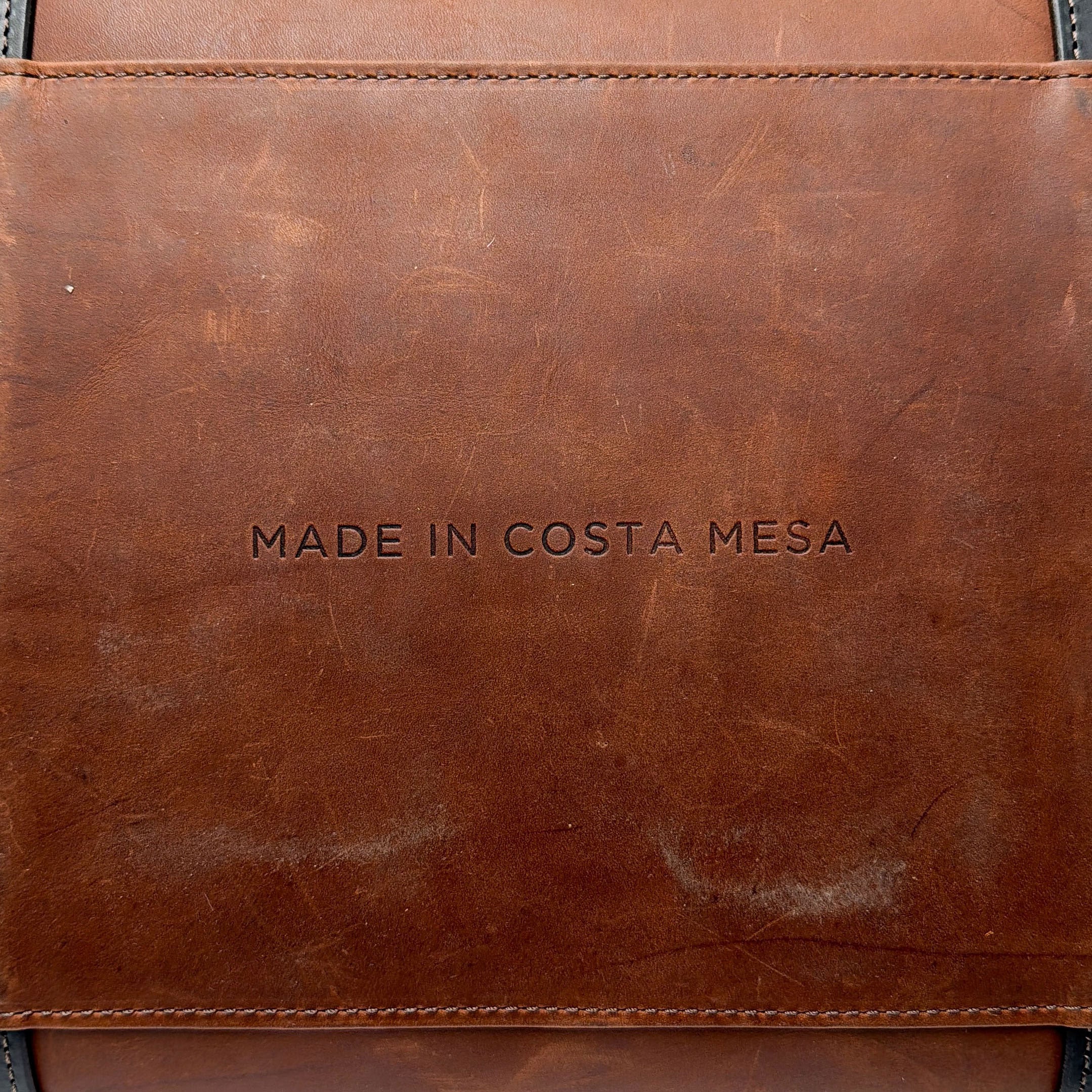 Brown leather duffle bag with MADE IN COSTA MESA text embossed on it.