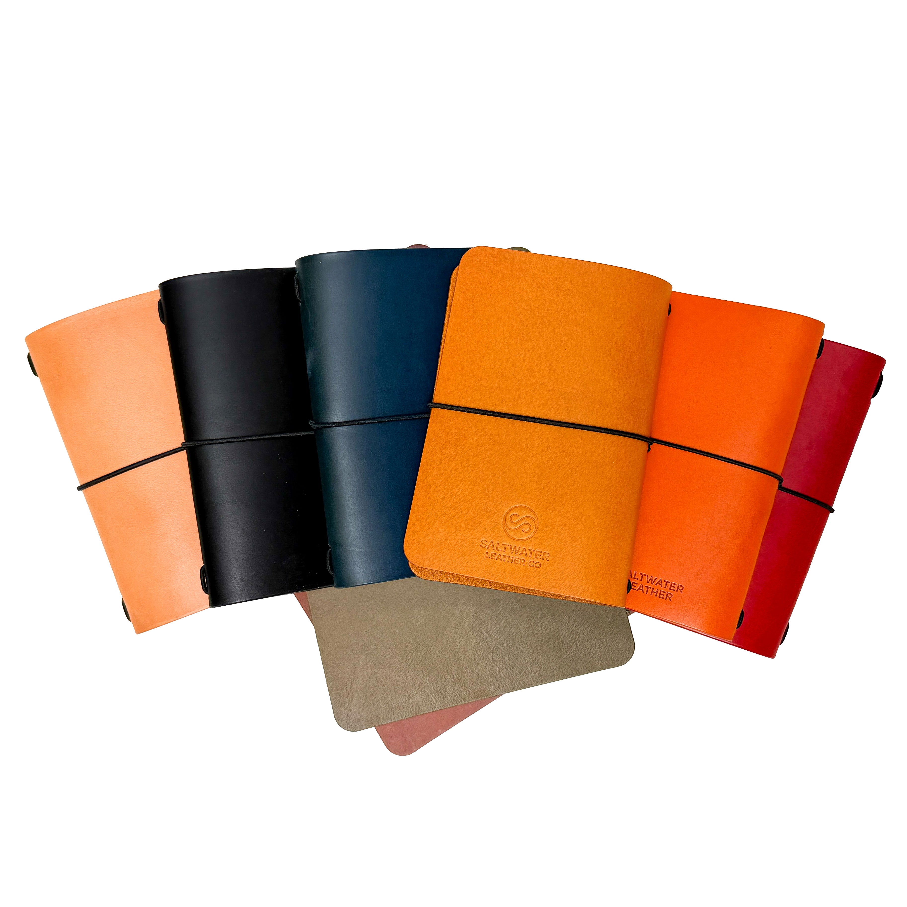 Saltwater Leather journal covers in six colors including California Poppy, Midnight, Pacific, Mustard, Cherry, and natural tan, fanned out against a white background with elastic band closures.