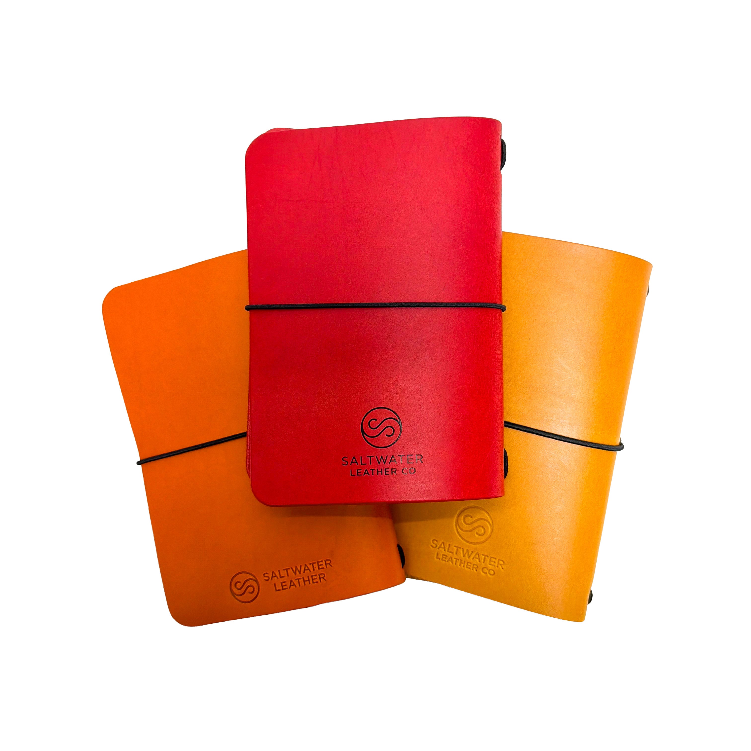 Three Saltwater Leather vegetable tanned leather journal covers in Cherry red, California Poppy orange, and Mustard yellow, fanned out against a white background with elastic band closures and embossed logo.